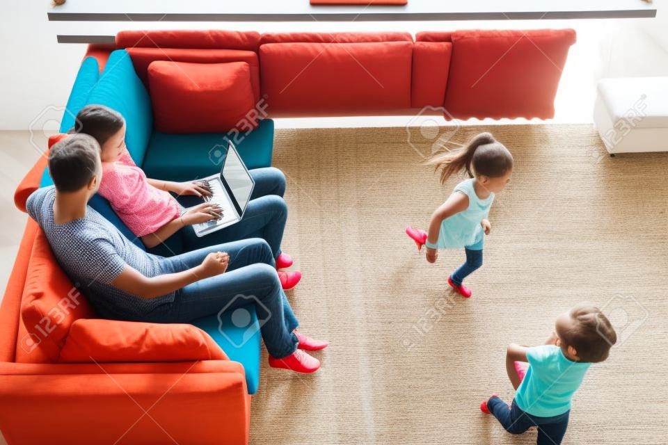Les petits enfants jouent au rattrapage pendant que les parents utilisent un ordinateur portable, sont assis sur un canapé, la mère et le père regardent une vidéo, naviguent, réservent des billets, voyagent, passent du temps en famille ensemble. Vue de dessus