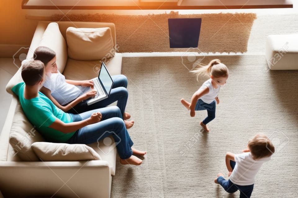 Les petits enfants jouent au rattrapage pendant que les parents utilisent un ordinateur portable, sont assis sur un canapé, la mère et le père regardent une vidéo, naviguent, réservent des billets, voyagent, passent du temps en famille ensemble. Vue de dessus