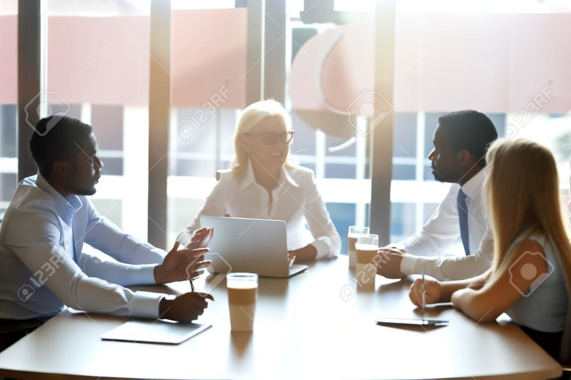 Five successful positive businesswomen and businessmen different age and ethnicity sitting in cozy boardroom office room and negotiating. Staff led by company boss gathered together starting briefing