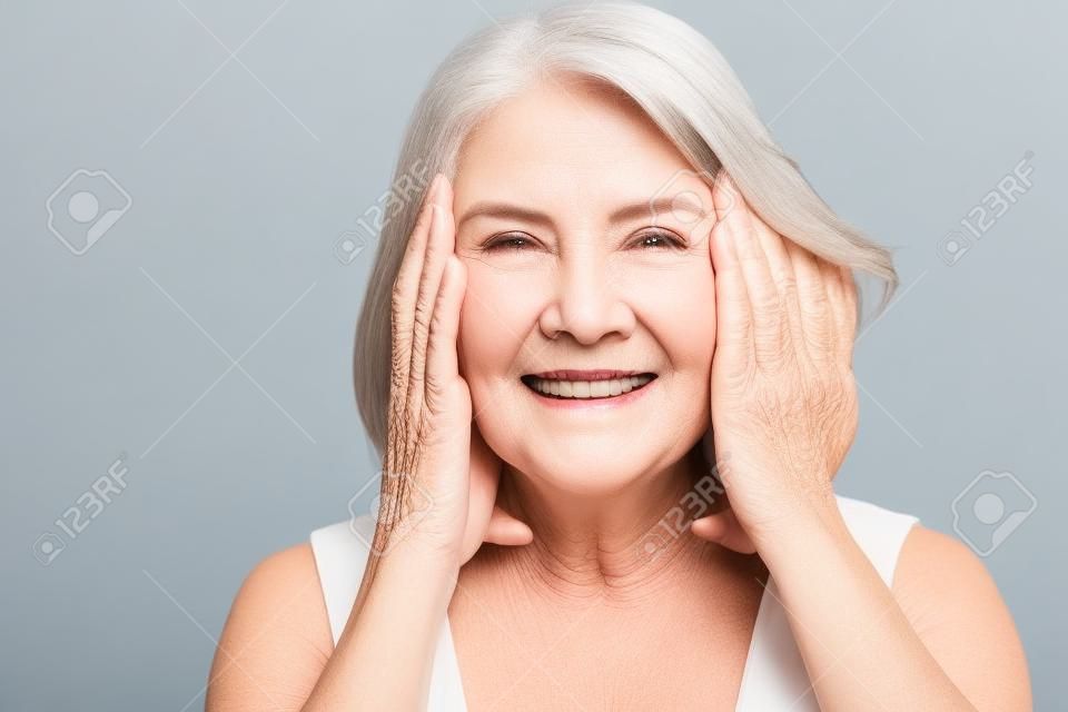 Happy mature older woman touching face healthy soft moisturized hydrated skin isolated on grey studio background, smiling middle aged senior lady natural beauty treatment anti aging concept, portrait