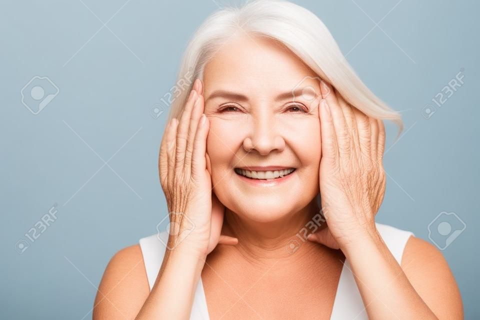 Gelukkig volwassen oudere vrouw aanraken gezicht gezonde zachte gehydrateerde huid geïsoleerd op grijze studio-achtergrond, glimlachende middelbare leeftijd senior dame natuurlijke schoonheid behandeling anti veroudering concept, portret