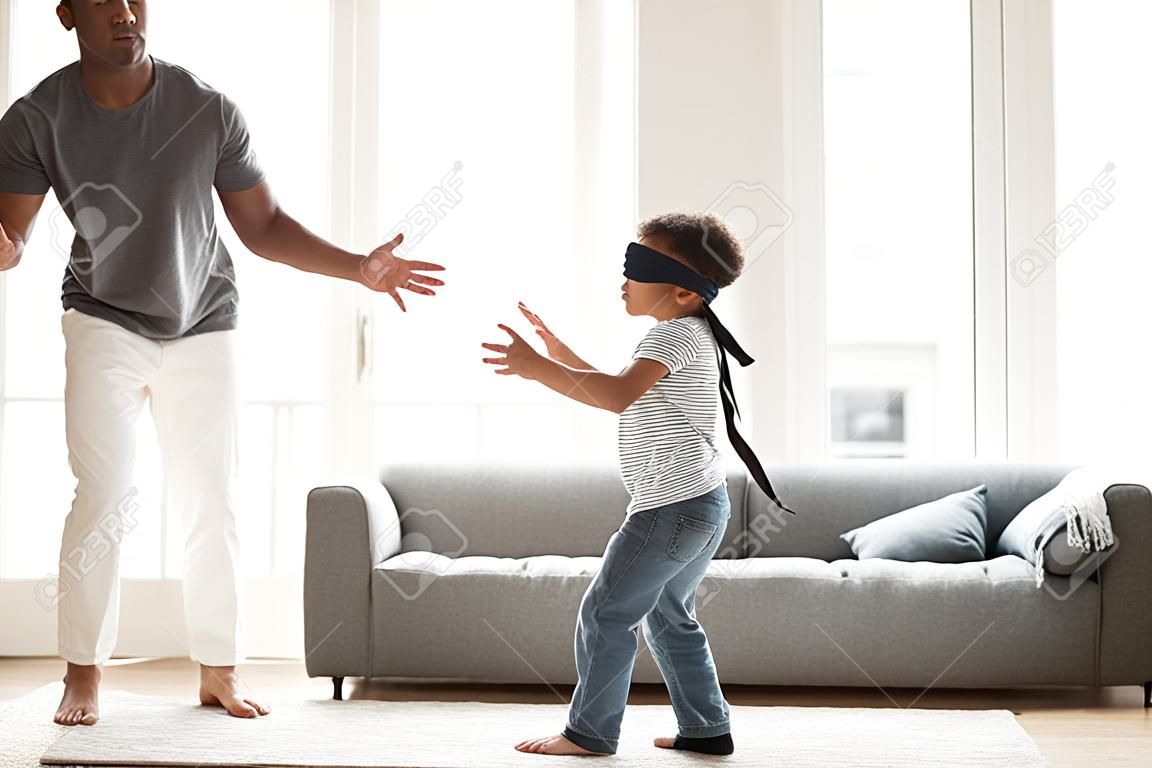 Funny little blindfolded boy play hide and seek game with mixed race family in living room, toddler have fun with black parents and sister, spend time together laughing entertaining at home