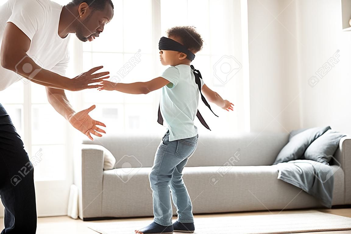 Funny little blindfolded boy play hide and seek game with mixed race family in living room, toddler have fun with black parents and sister, spend time together laughing entertaining at home
