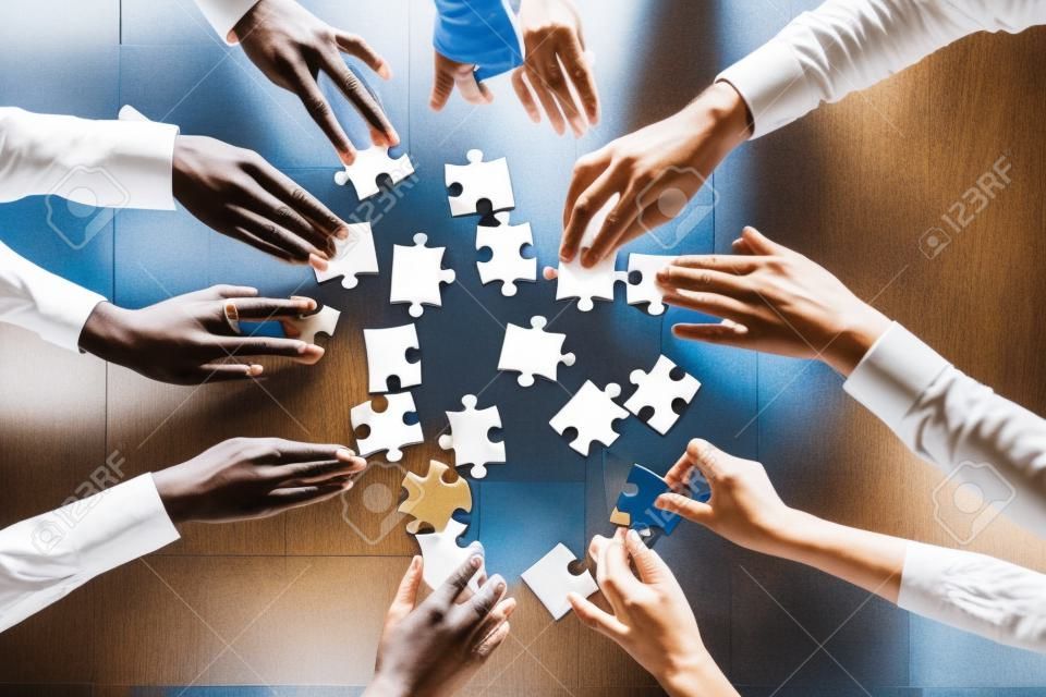 Diverse business team people hands assemble puzzle together connect pieces at desk, employees collaborate find common solution engaged help contribute in effective teamwork concept top close up view