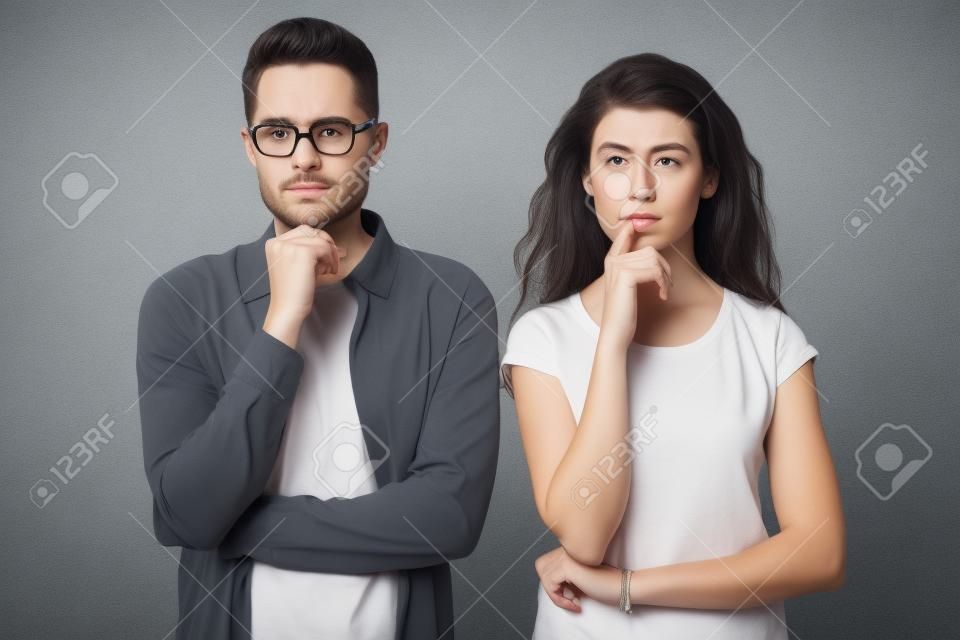 Pensive millennial couple stand isolated on grey studio background touch chin with finger thinking of something, thoughtful man and woman consider or plan idea, look at camera lost in thoughts