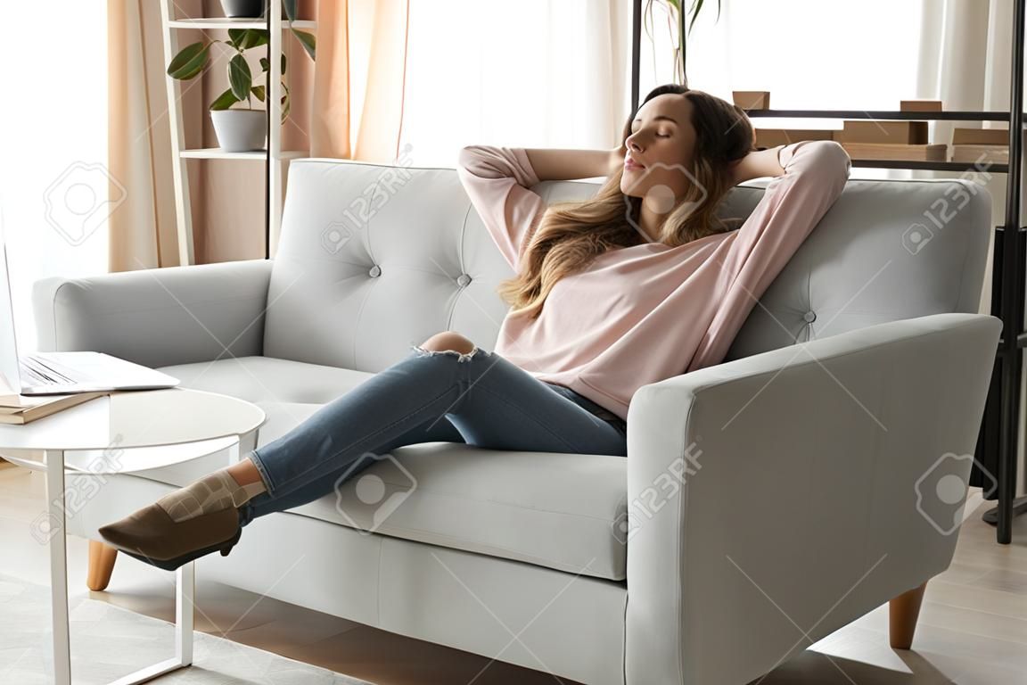 Relaxed young woman leaned on couch closed eyes putting hands behind head enjoy fresh air, freelancer resting from work in modern cozy living room alone, daydream day nap, fall asleep or pause concept