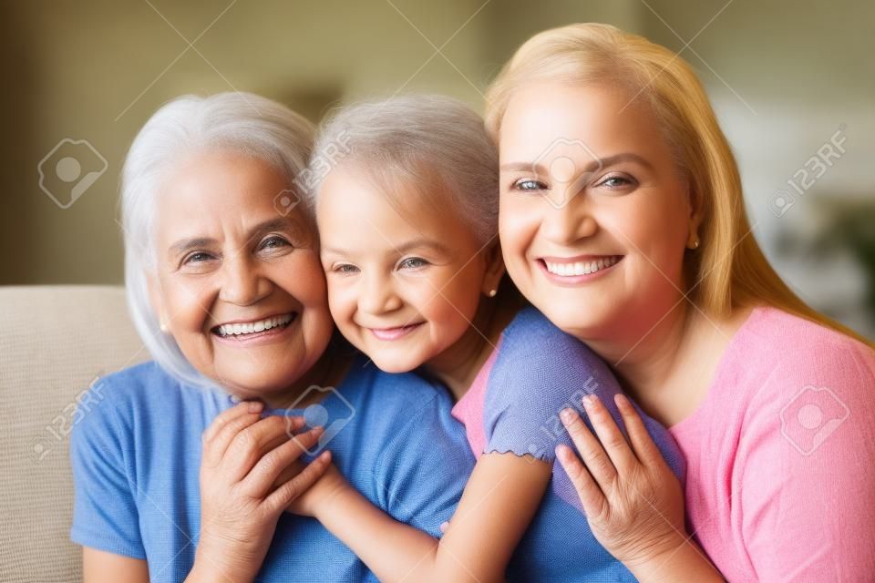 Head shot of three diverse generations indoors. Portrait of beautiful little granddaughter daughter and grandmother smiling looking at camera sitting together on couch hugging in living room at home