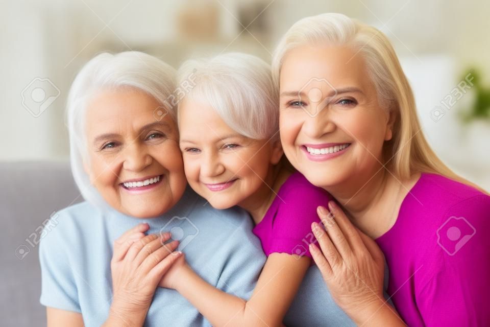 Head shot of three diverse generations indoors. Portrait of beautiful little granddaughter daughter and grandmother smiling looking at camera sitting together on couch hugging in living room at home
