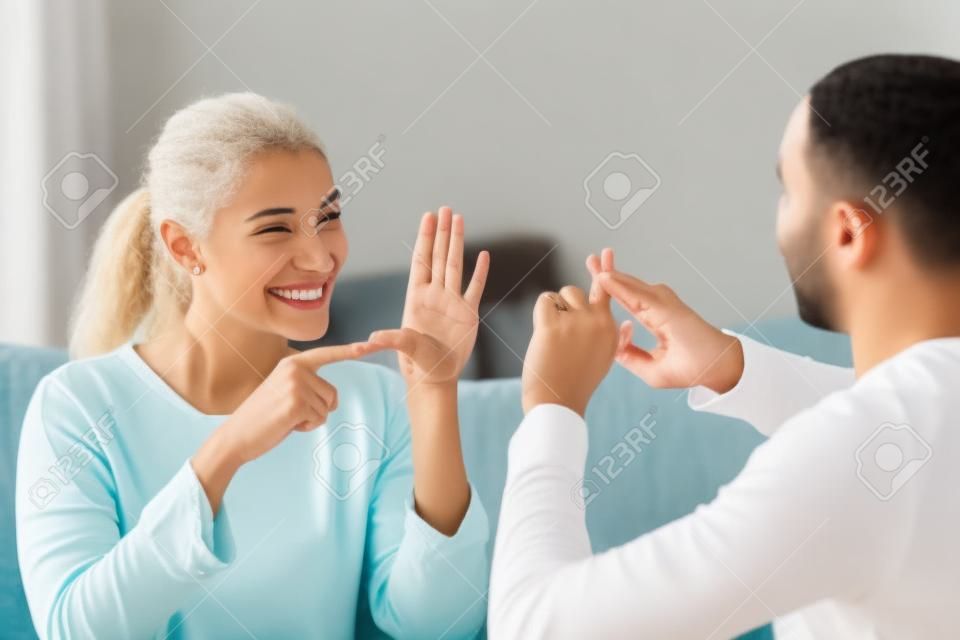Un couple souriant d'ethnies mixtes ou des amis interraciaux parlant avec le langage de la main du doigt des signes, heureux deux sourds et muets malentendants communiquant à la maison assis sur un canapé montrant des gestes de la main