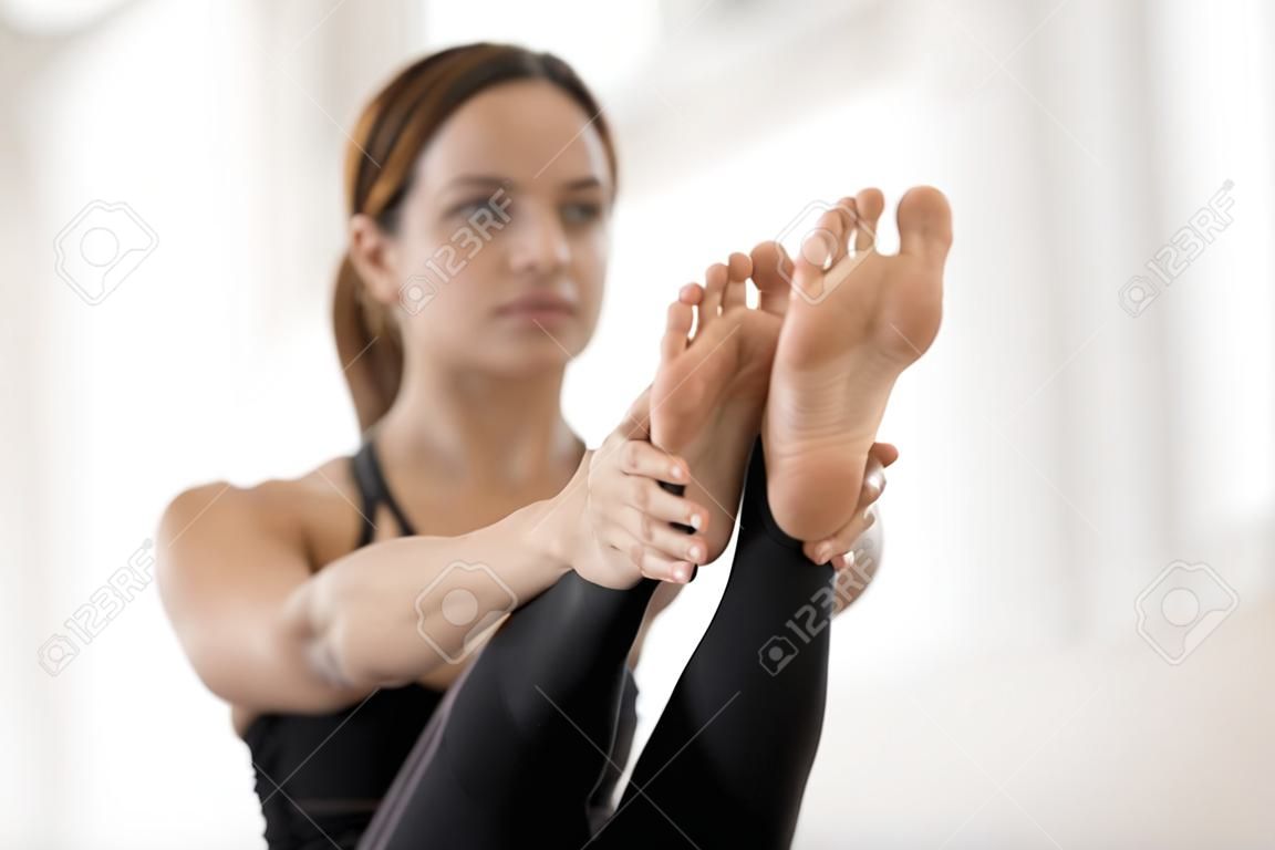 Beautiful young woman doing Paripurna Navasana exercise, boat pose, practicing yoga, feet close up, attractive sporty girl wearing black sportswear working out at home or in yoga studio