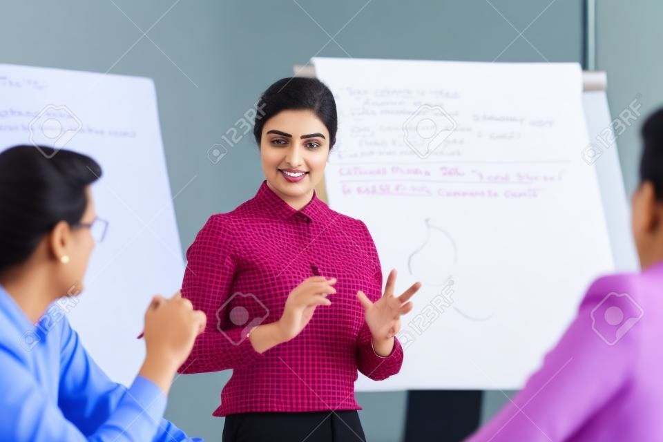 Confident female indian conference speaker coach training diverse employees group give flip chart presentation at office meeting, hindu business woman presenter teach staff team at corporate workshop
