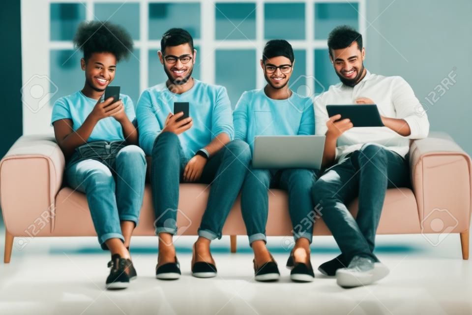 Diverse young people using electronic devices, phones, laptop, tablet, sitting in row on couch, obsessed by gadgets, chatting in social network, watching video, playing games, ignoring each other