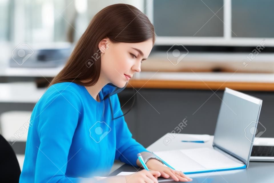 La ragazza caucasica concentrata si siede alla scrivania studiando la preparazione per l'esame della sessione universitaria tenendo la penna che scrive nel libro di testo, studentessa intelligente focalizzata che prende appunti facendo attività usando il laptop al chiuso