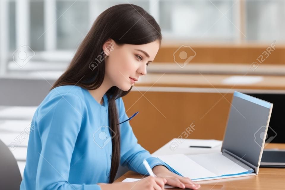 Konzentriertes kaukasisches Mädchen sitzt am Schreibtisch und lernt sich auf die Prüfung der Universitätssitzung vorbereiten und hält den Stift im Lehrbuch nieder