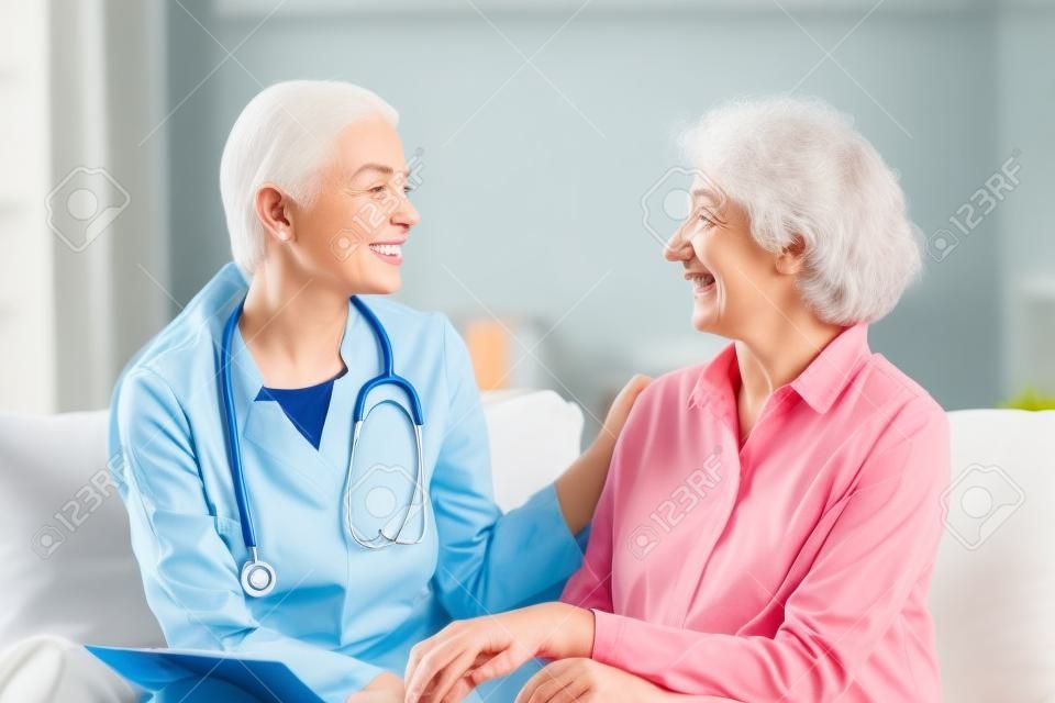 A enfermeira jovem feliz fornece o serviço médico da assistência médica ajuda a apoiar a avó velha sorridente na visita médica do cuidador homecare, doutor da senhora dá a empatia encoraje o paciente aposentado senta-se no sofá no hospital home