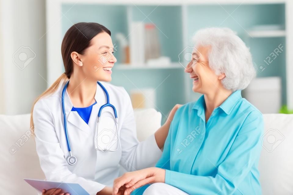幸せな若い女性看護師は、ホームケアの医療訪問で笑顔の古いおばあちゃんをサポートするケア医療サービスを提供し、女性の介護者の医師は、自宅の病院でソファに座って引退した患者を奨励する共感を与えます
