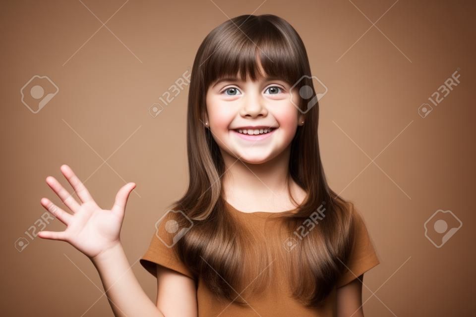 Close up head shot portrait with funny attractive little brown-haired girl. Concept happy activity playful kid gives welcome gesture on gray background, six year child looking at camera and posing.