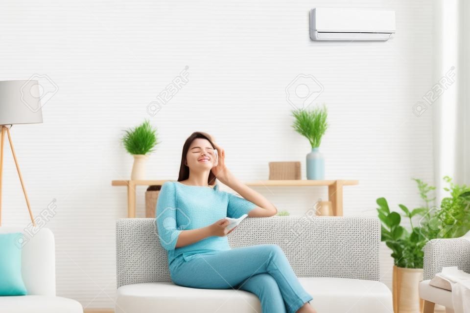 50s woman rest on couch closed eyes enjoy fresh air hold remote control use air conditioner cools herself at summer hot day adjusting temperature inside of living room, comfort wellbeing life concept