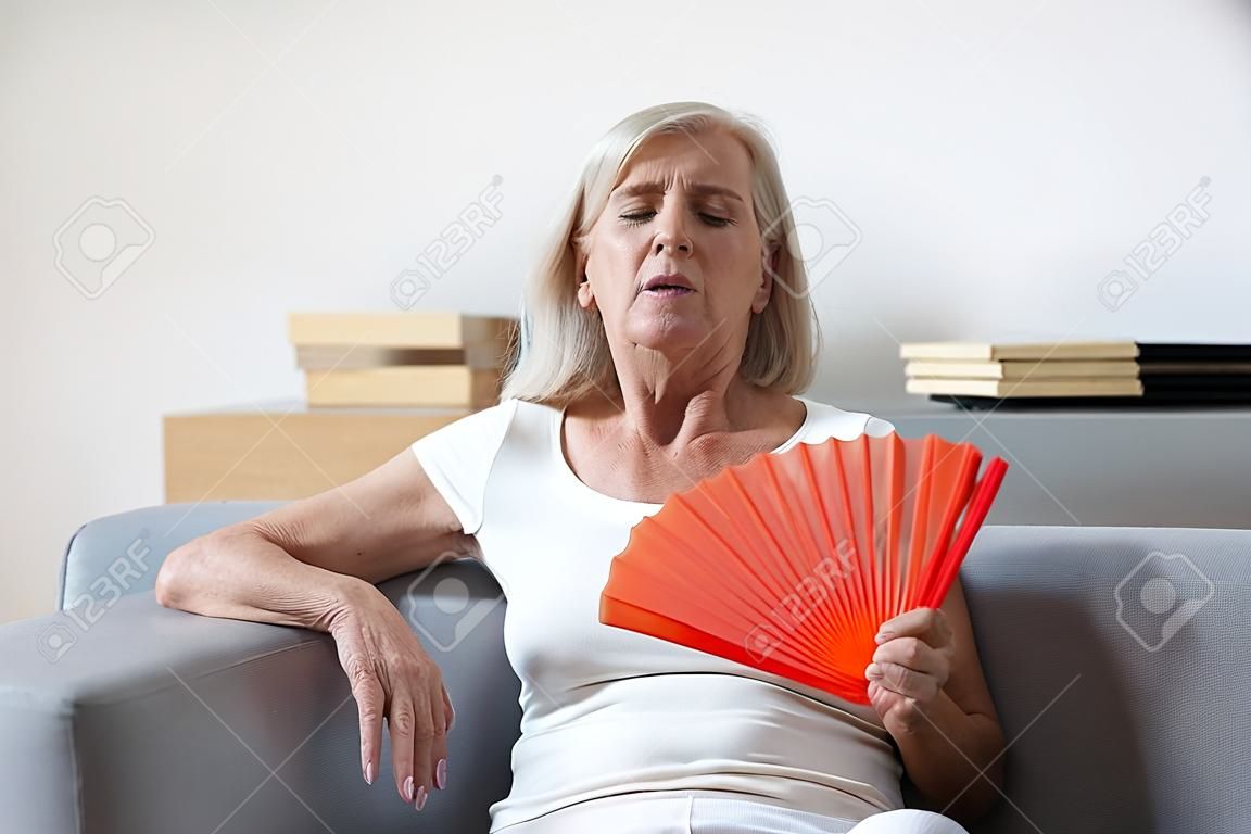 Unwell elderly female retiree sit on couch feel dehydrated tired of heat use hand waver to cool down