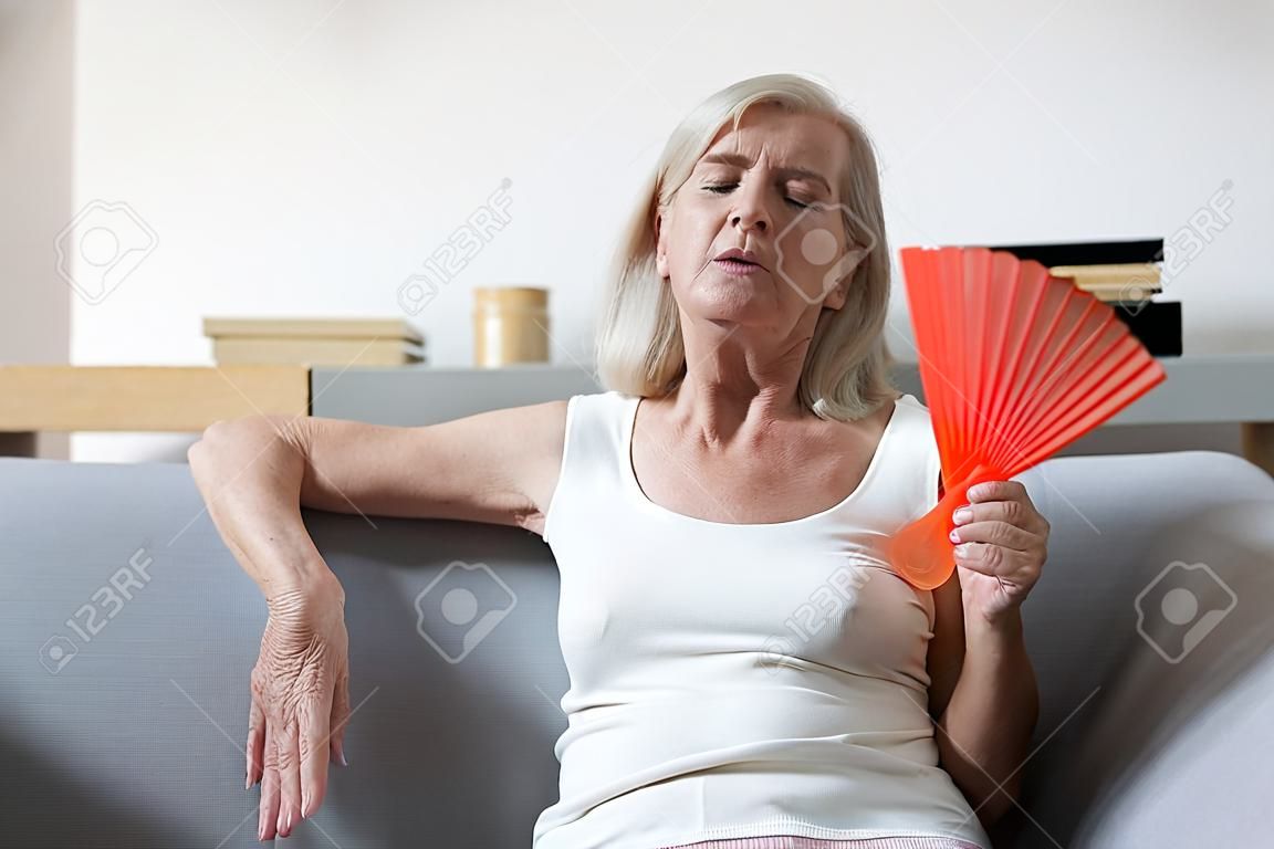 Unwell elderly female retiree sit on couch feel dehydrated tired of heat use hand waver to cool down