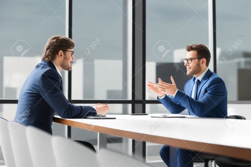 Male colleagues sit at office desk talk discuss company project ideas at briefing together, concentrated man business partners speak negotiate brainstorm at company meeting in boardroom