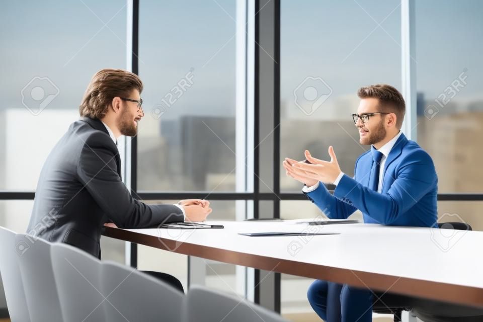 Male colleagues sit at office desk talk discuss company project ideas at briefing together, concentrated man business partners speak negotiate brainstorm at company meeting in boardroom