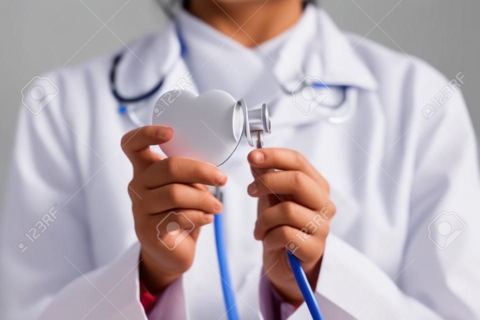 Close up young indian female doctor in white uniform putting stethoscope to heart figure, advertising professional cardiology services, preventing cardiovascular diseases or giving consultation.
