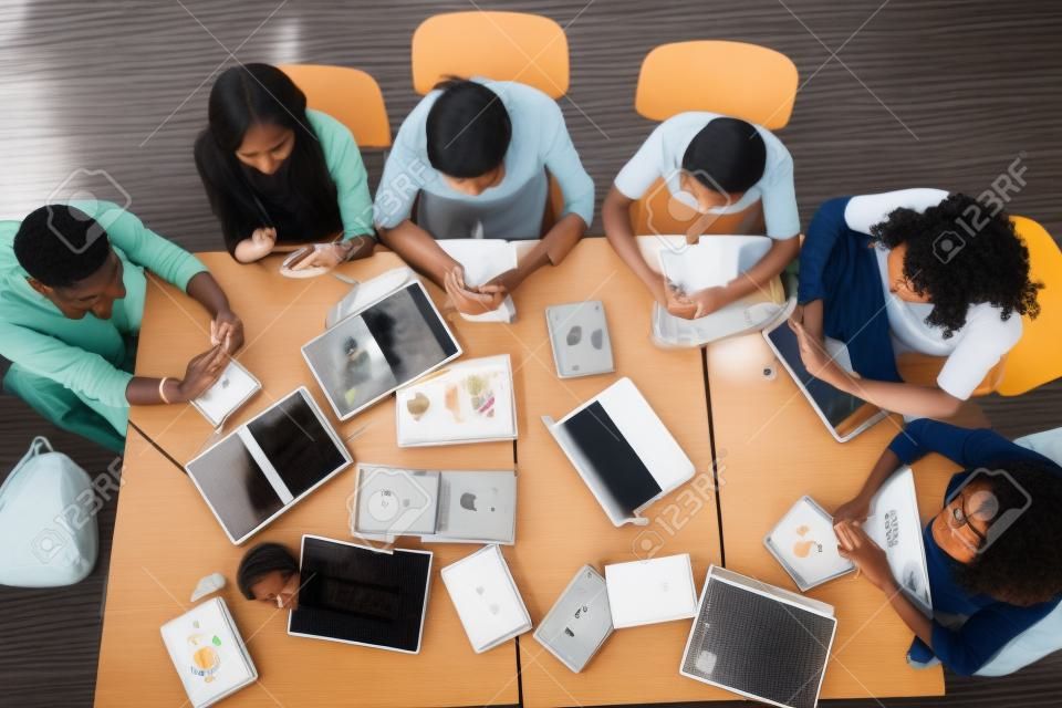 Grupa wieloetnicznych uczniów z widokiem z góry korzysta ze smartfonów siedzących przy wspólnym stole w klasie. korzystanie z aplikacji mobilnych w przerwie, nadużywanie nowoczesnej technologii bezprzewodowej przez młodych ludzi, zły nawyk, koncepcja stylu życia