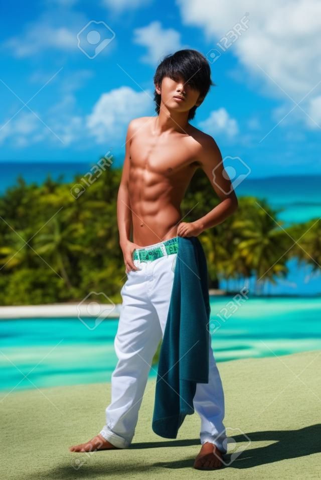 Teenage Asian skinny shirtless boy striking a fashion pose in a tropical landscape with bay in the background.