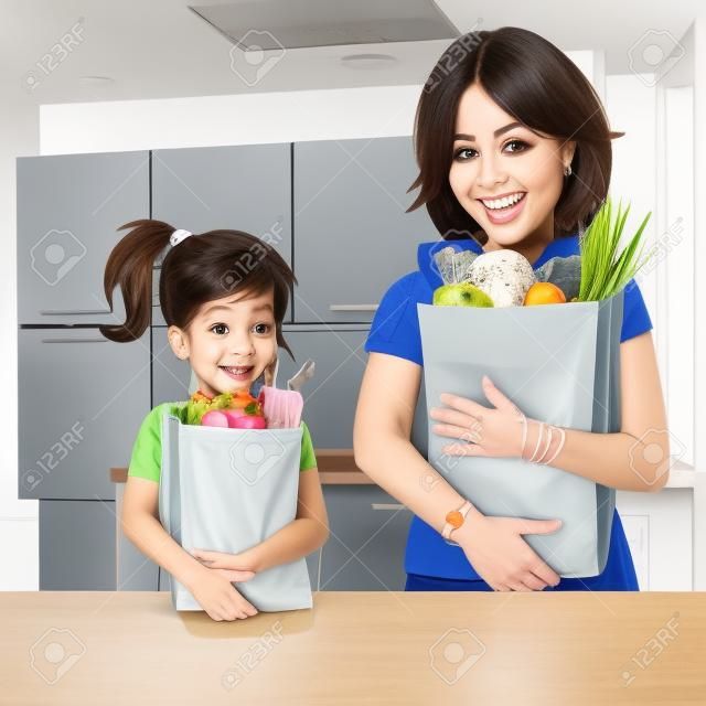 cute young daughter helps her mother to carry groceries paper bags in kitchen