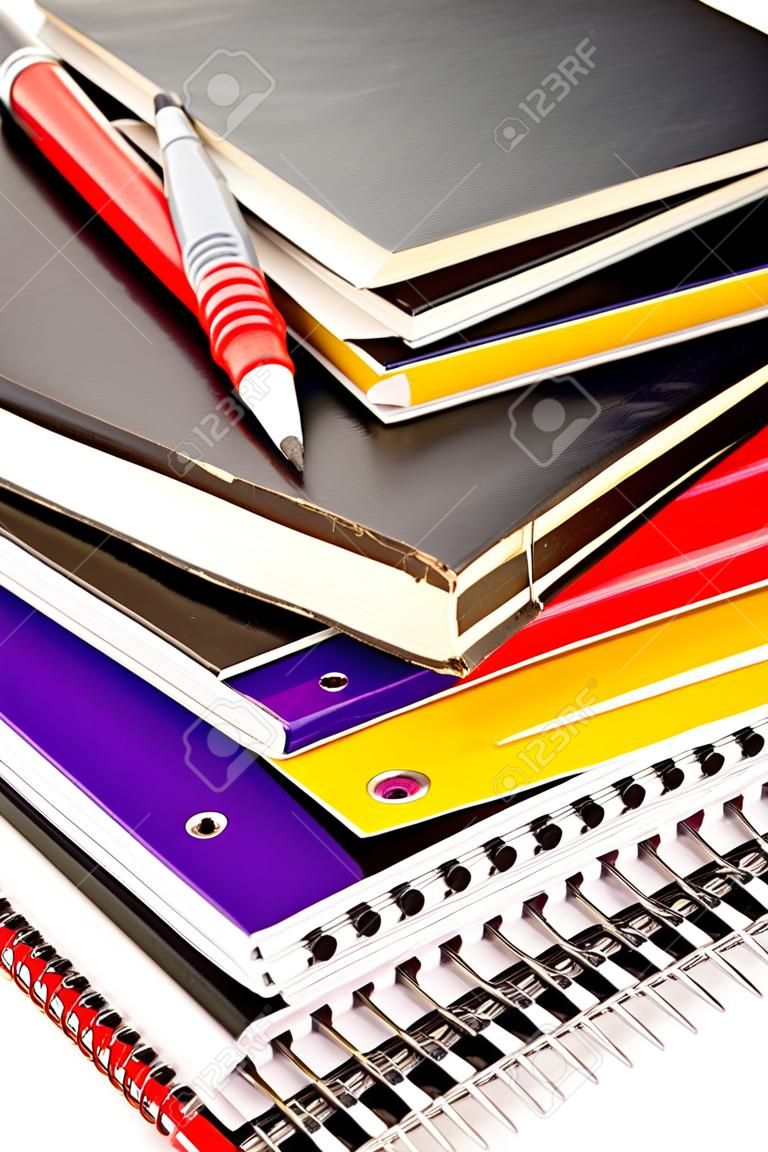 A stack of school books and spiral notebooks with a pencil and pens on tops in front of a white background