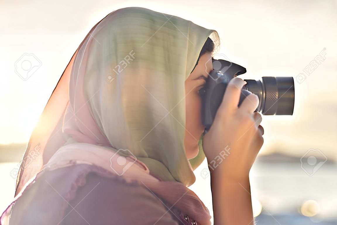 Fotógrafo árabe da mulher em um lenço que toma a foto usando a câmera no fundo do por do sol.