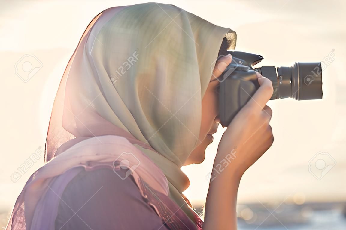 Fotógrafo árabe da mulher em um lenço que toma a foto usando a câmera no fundo do por do sol.