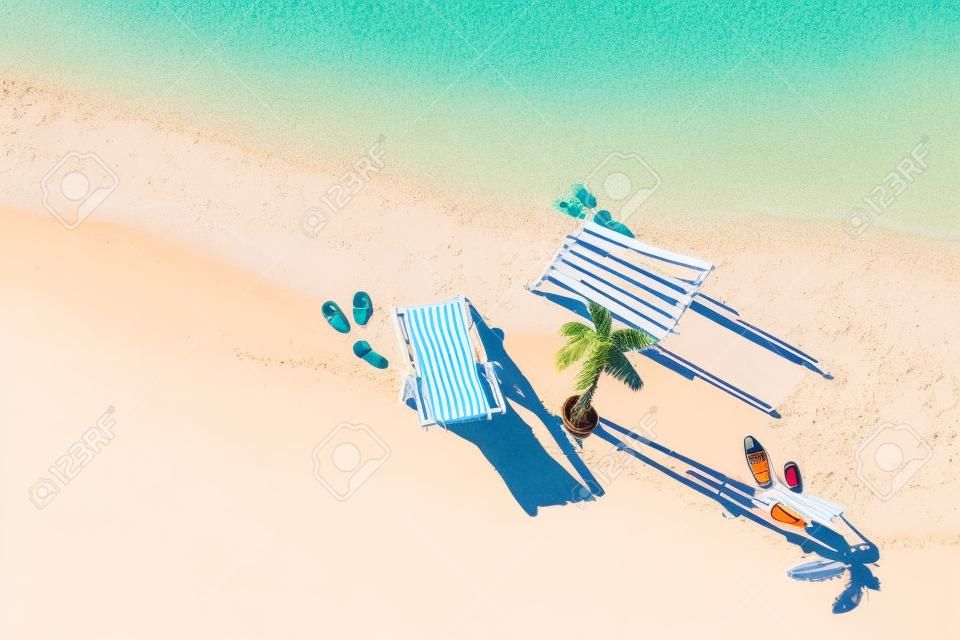 Aerial view of two deck chair, sunbed, lounge, glass of orange juice, flip flops, palm tree on sandy beach. Summer and travel concept. Minimalism