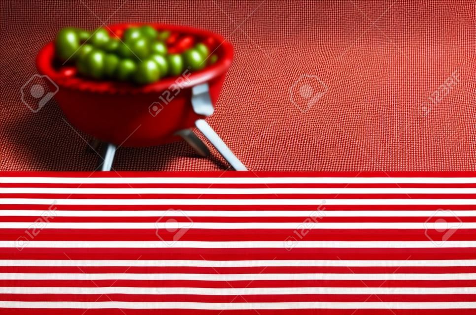 Lege picknick tafel achtergrond bedekt met een vers land rood en wit gecontroleerd doek voor uw product plaatsing of reclame met een barbecue op een groene gazon achter
