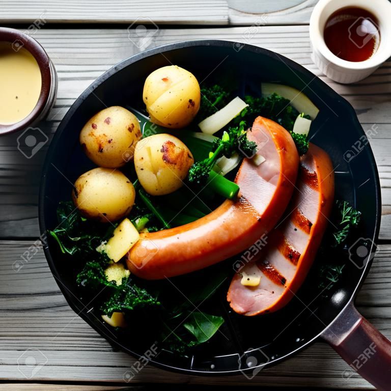 Close up Aerial Shot of Gourmet German Recipe with Sausage, Pork and Potatoes with Kale on a Frying Pan. Served on Wooden Table with Mustard Sauce and Beer on the Side.