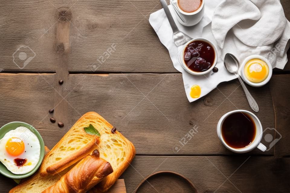 Le petit-déjeuner se répand avec du café, du pain, des croissants frais, des œufs frits, du miel et des conserves sur une ancienne table rustique en bois vert, vue aérienne avec copyspace