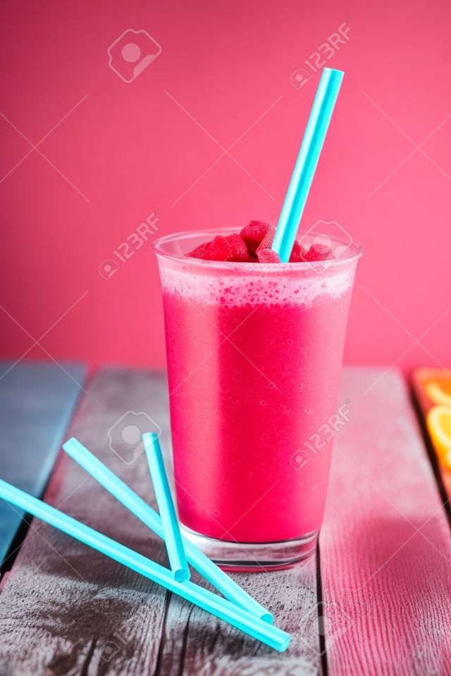 Still Life Profile of Refreshing and Cool Frozen Red Fruit Slush Drink in Plastic Cup Served on Rustic Wooden Table with Collection of Colorful Straws