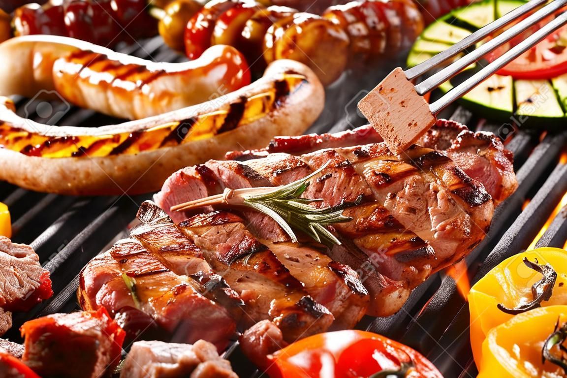 Köstliche Auswahl an Fleisch Nackensteak und Gemüse Grillen auf einem Grill mit Würstchen aus Schweinefleisch, Koteletts, Spieße mit gemischten Kebab, Paprika und Auberginen in einer Nahaufnahme Ansicht mit einer Zange ein Kotelett drehen