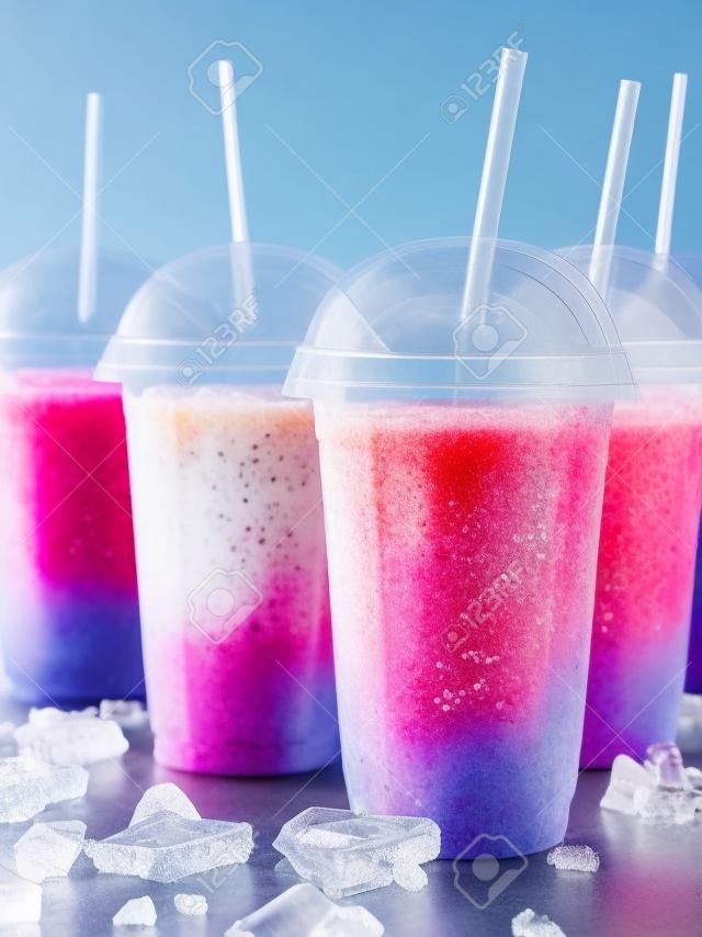 Still Life Profile of Frozen Fruit Slush Granita Drinks in Plastic Take Away Cups with Lids and Drinking Straws Chilling on Cold Metal Surface with Scattered Ice Cubes