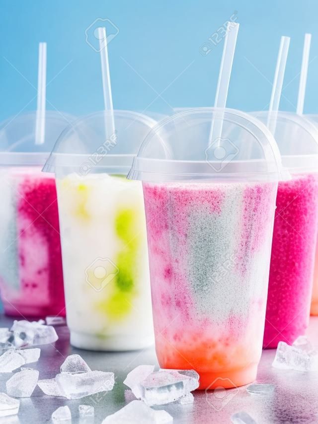 Still Life Profile of Frozen Fruit Slush Granita Drinks in Plastic Take Away Cups with Lids and Drinking Straws Chilling on Cold Metal Surface with Scattered Ice Cubes