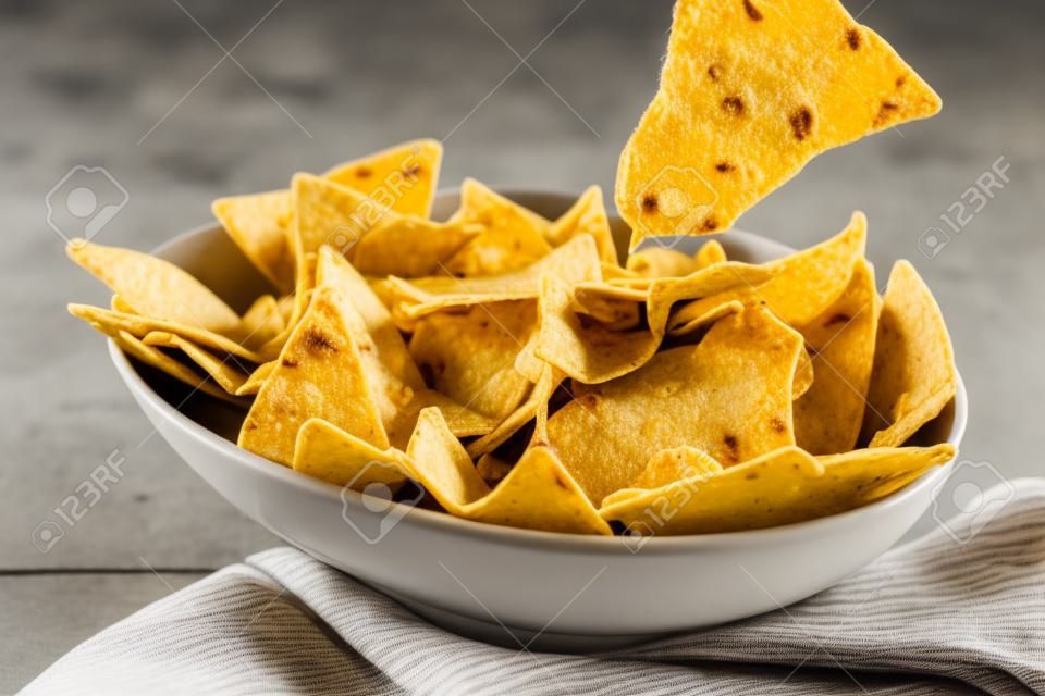 Single triangular yellow corn tortilla chip pulled out of bowl of cheese covered nachos over gray and white tablecloth