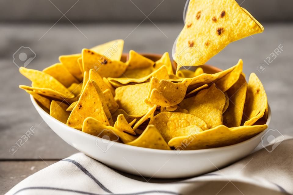 Single triangular yellow corn tortilla chip pulled out of bowl of cheese covered nachos over gray and white tablecloth