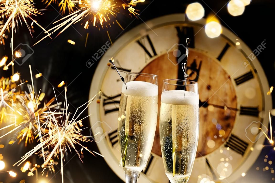 Festive New Years background with sparklers and champagne in front of a clock counting down to midnight with sparkling bokeh on darkness
