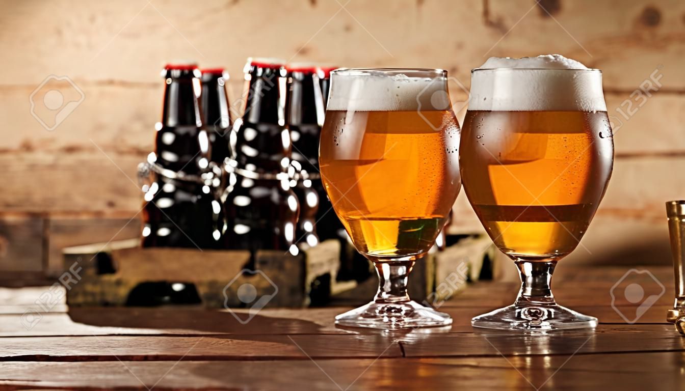 Two glasses of chilled beer with frothy heads standing together on a bar counter with a crate of beer bottles behind