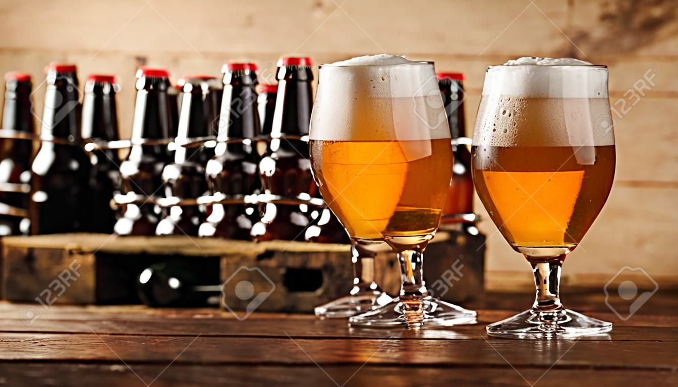 Two glasses of chilled beer with frothy heads standing together on a bar counter with a crate of beer bottles behind
