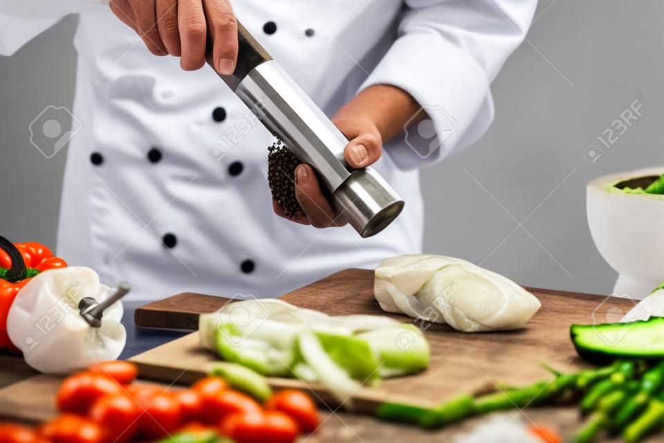 전경에서 야채에 소금에 절인 고기 그물에 대한 흰색 재킷 연삭 후추에 남성 요리사의 측면보기