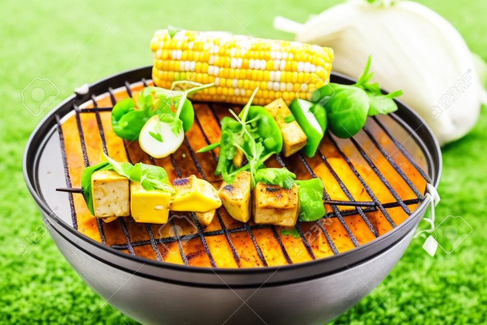 Groenten en tofu kebab grillen op rooster grille met verse maïs, gezien in close-up tegen groen gras op de achtergrond