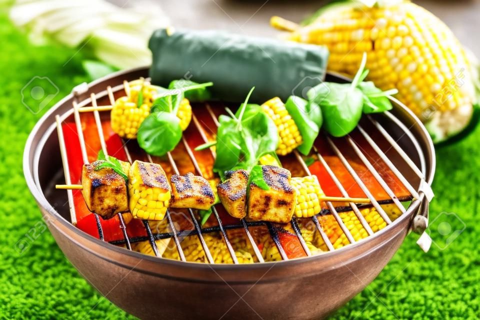 Verdure e tofu kebab grigliate sulla griglia con mais fresco, visto in primo piano contro il prato verde erba in background