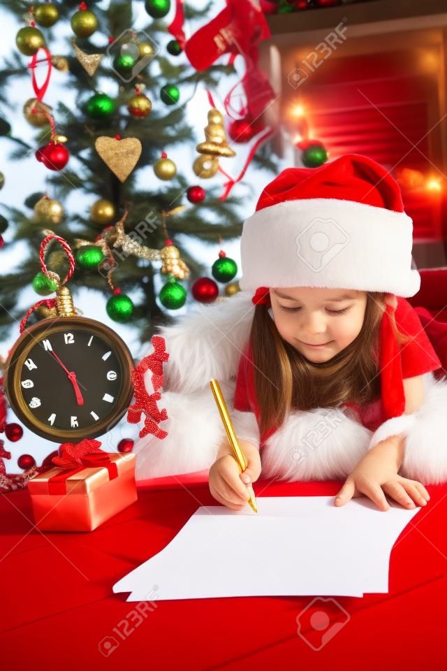 Funny girl in Santa hat writes letter to Santa near christmas tree and clock